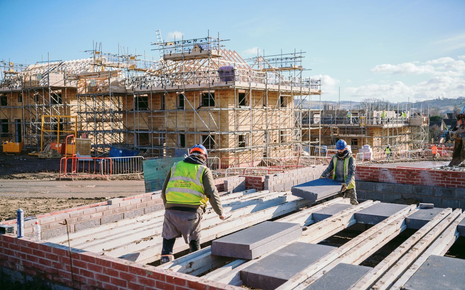 Two construction workers in high-visibility vests and hard hats installing grey insulation blocks between concrete floor beams on a new housing development site with scaffolding in the background.
