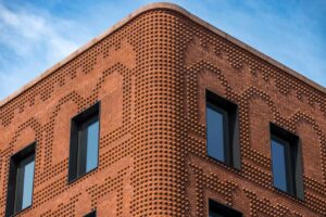 Close-up of a sculpted brick façade with protruding nodules forming a rhythmic texture, framed by black window openings under a clear blue sky.