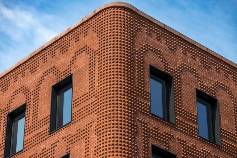 Close-up of a sculpted brick façade with protruding nodules forming a rhythmic texture, framed by black window openings under a clear blue sky.