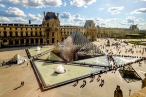 Aerial view of the Louvre Museum courtyard featuring I.M. Pei’s glass pyramid, surrounded by historic facades and reflecting pools under a partly cloudy sky.
