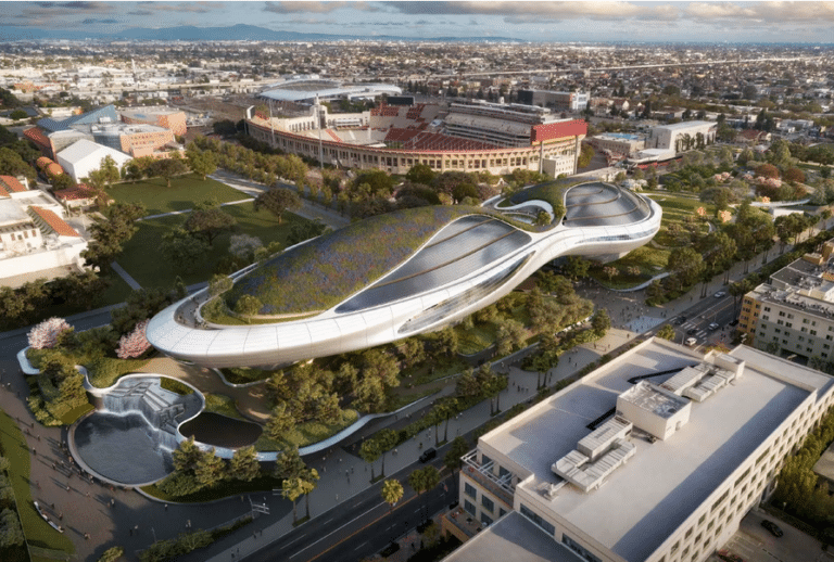 A futuristic, aerodynamic white building with a green rooftop garden, situated in a lush park next to the Los Angeles Memorial Coliseum.