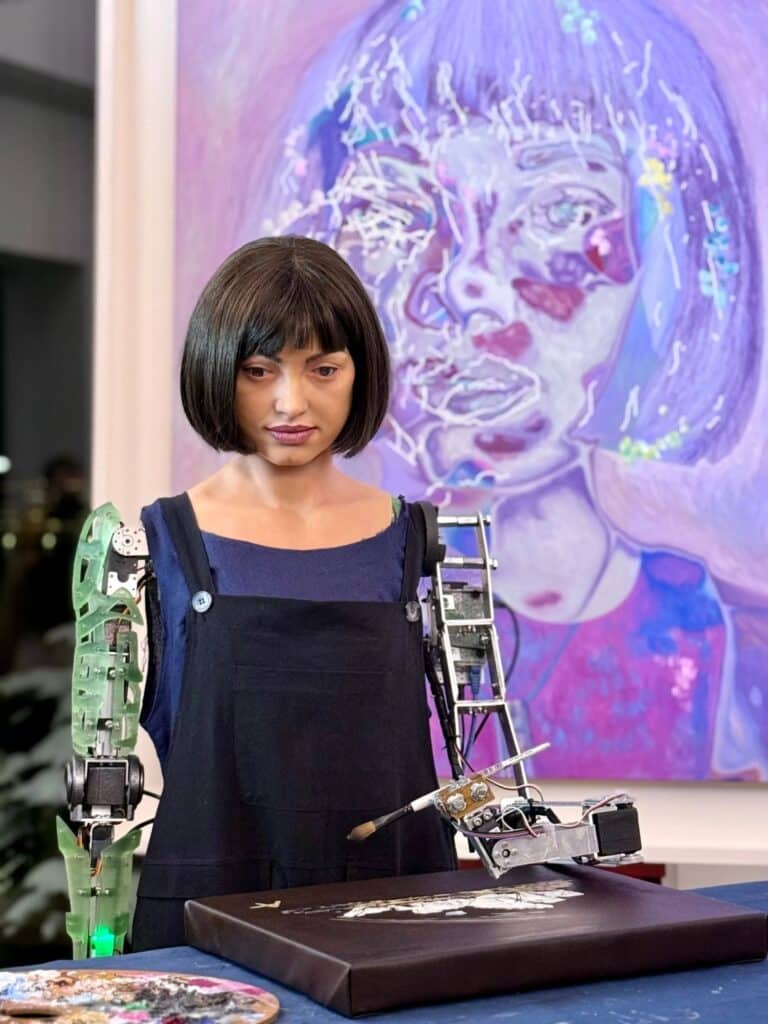 The humanoid robot artist Ai-Da is shown actively creating a piece of art with her robotic arm in a studio setting.