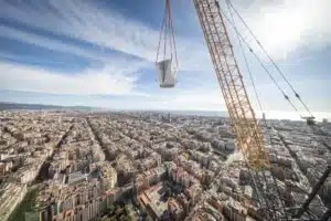 Yellow construction crane hoisting the final white architectural piece of the Sagrada Família tower against the Barcelona cityscape and Mediterranean horizon.