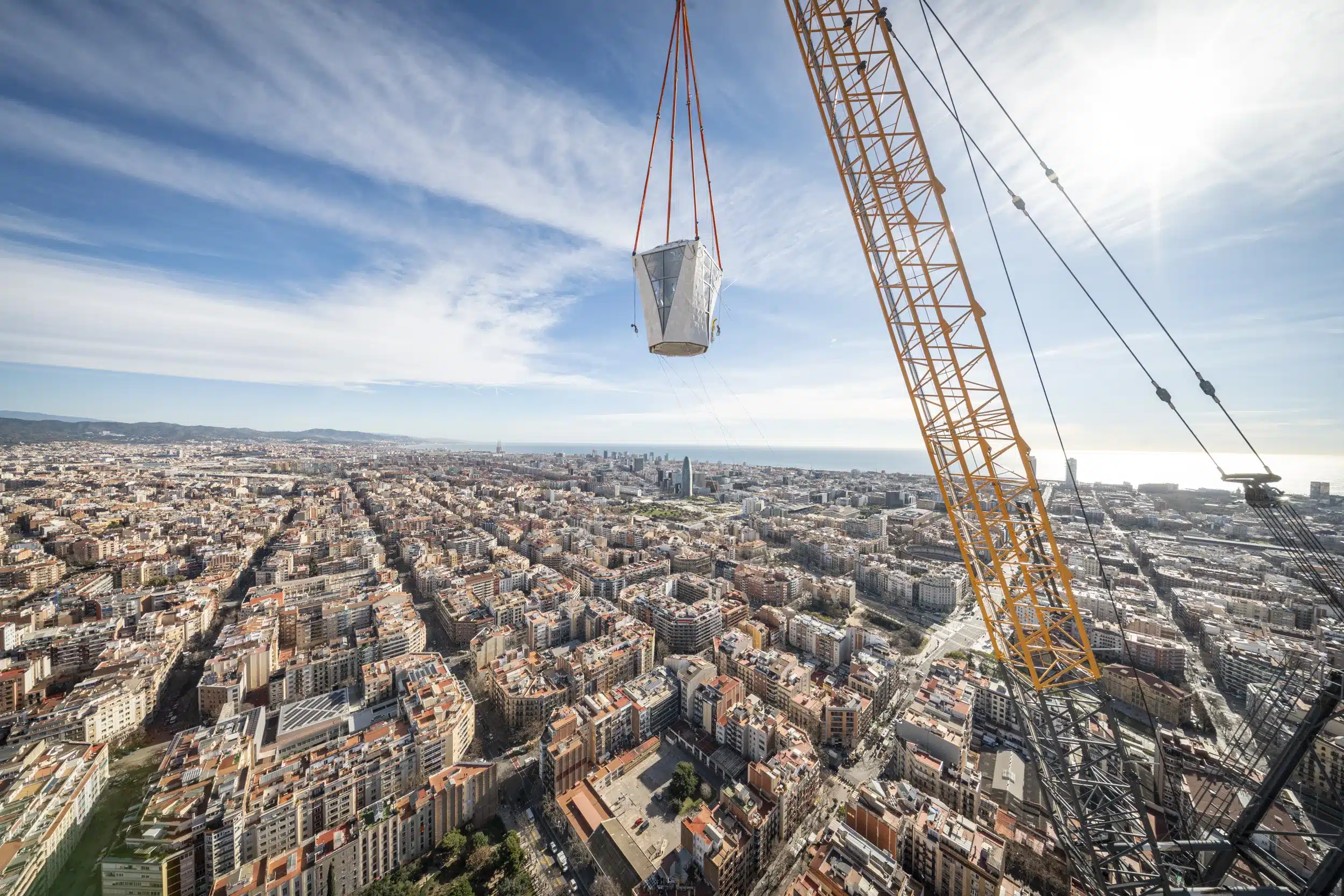 Yellow construction crane hoisting the final white architectural piece of the Sagrada Família tower against the Barcelona cityscape and Mediterranean horizon.