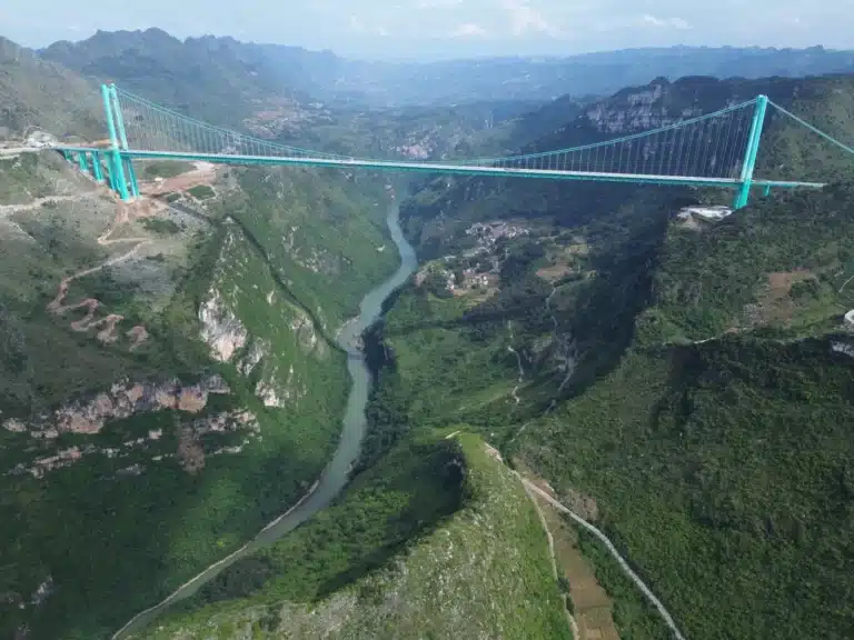 Panoramic aerial view of the Huajiang Canyon Bridge suspension structure spanning a deep green gorge with the Beipan River winding below.