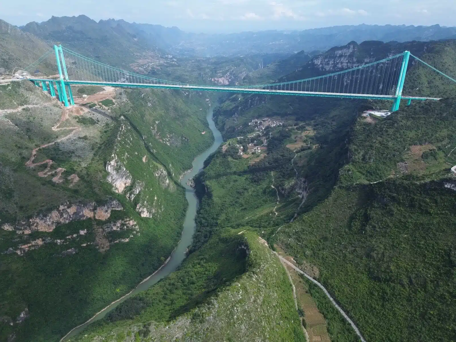 Panoramic aerial view of the Huajiang Canyon Bridge suspension structure spanning a deep green gorge with the Beipan River winding below.