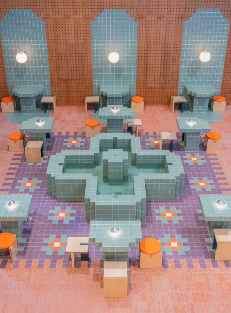 High-angle view of a central cross-shaped water fountain clad in pixelated teal tiles, surrounded by geometric purple flooring, custom tiled tables, and orange stools within a wooden-paneled room.