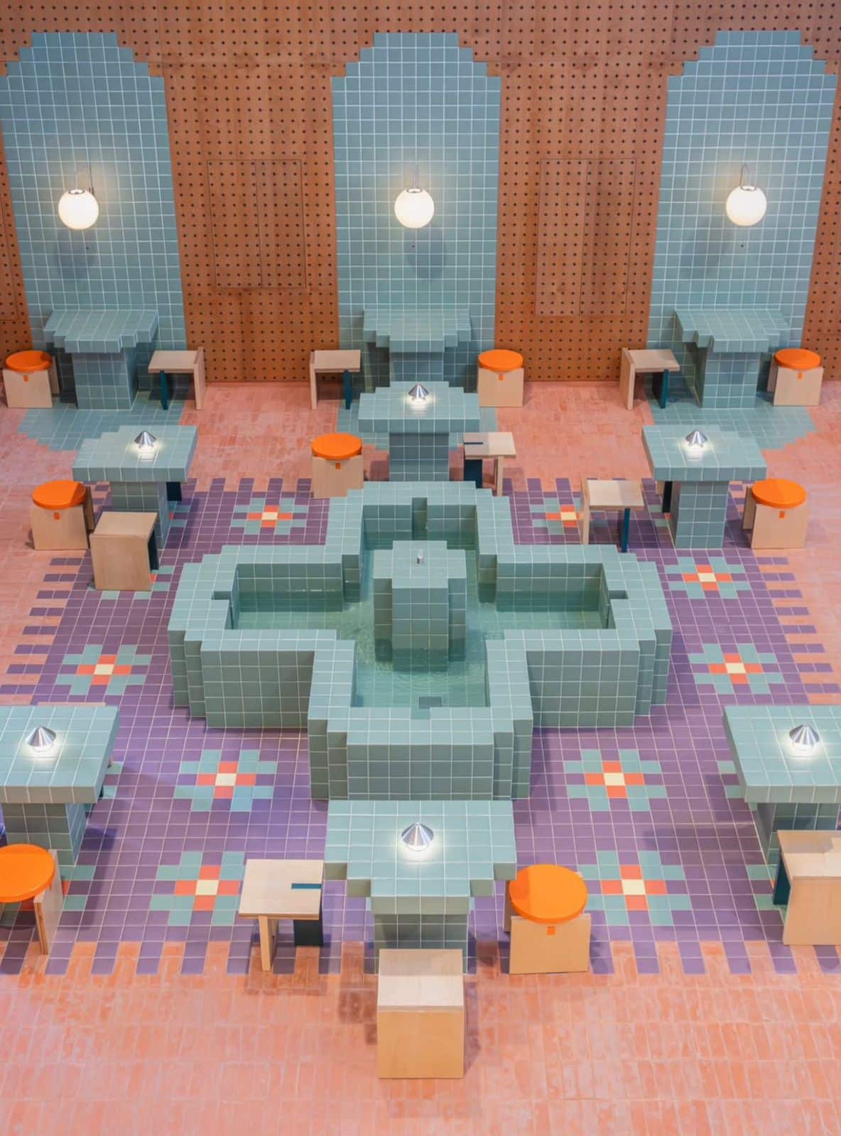 High-angle view of a central cross-shaped water fountain clad in pixelated teal tiles, surrounded by geometric purple flooring, custom tiled tables, and orange stools within a wooden-paneled room.