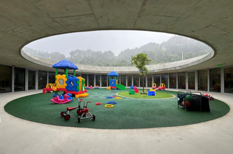 View from under the concrete overhang of the circular school courtyard featuring a playground during light rain.