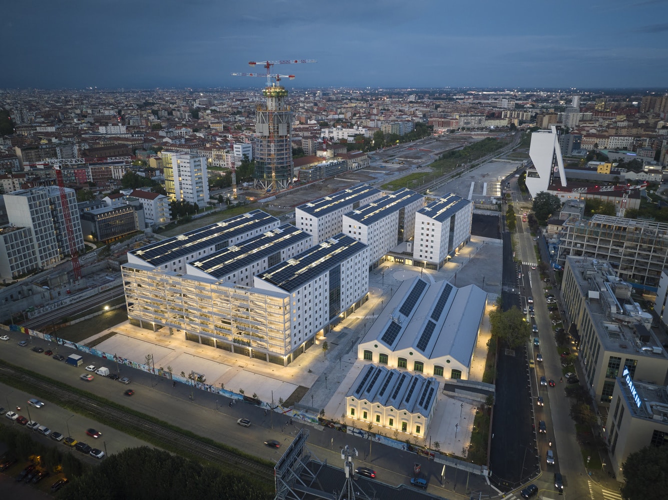 An aerial view of the sustainable Milano Cortina 2026 Olympic Village at dusk, showing modern residential buildings with solar panels and the surrounding Milan cityscape.