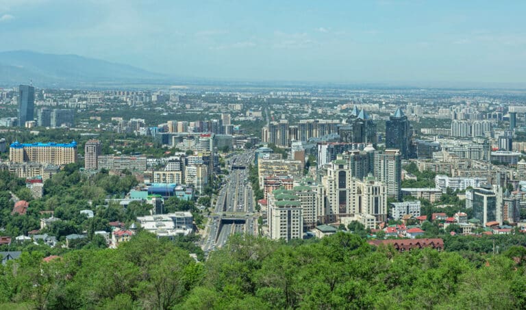 High-angle view of the modern Almaty architecture featuring the glass-clad Esentai Tower and Al-Farabi Avenue against a hazy mountain backdrop.