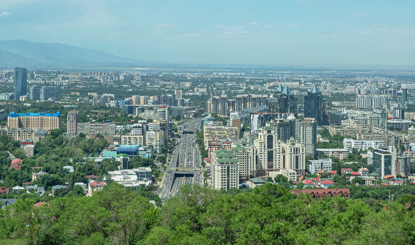 High-angle view of the modern Almaty architecture featuring the glass-clad Esentai Tower and Al-Farabi Avenue against a hazy mountain backdrop.
