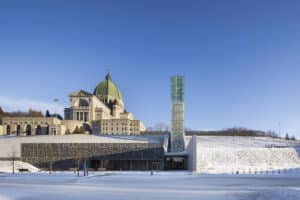 A towering glass and wood bell tower rises above stone gabion walls at the Montreal Oratory Pavilion.