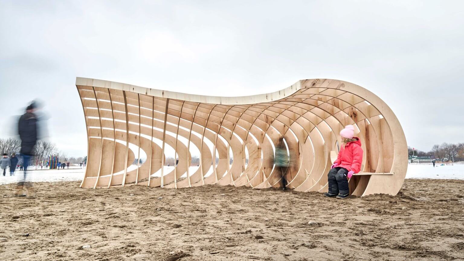 Curved plywood structure with waffled ribs resembling a crashing wave on a snowy beach at Winter Stations Toronto.