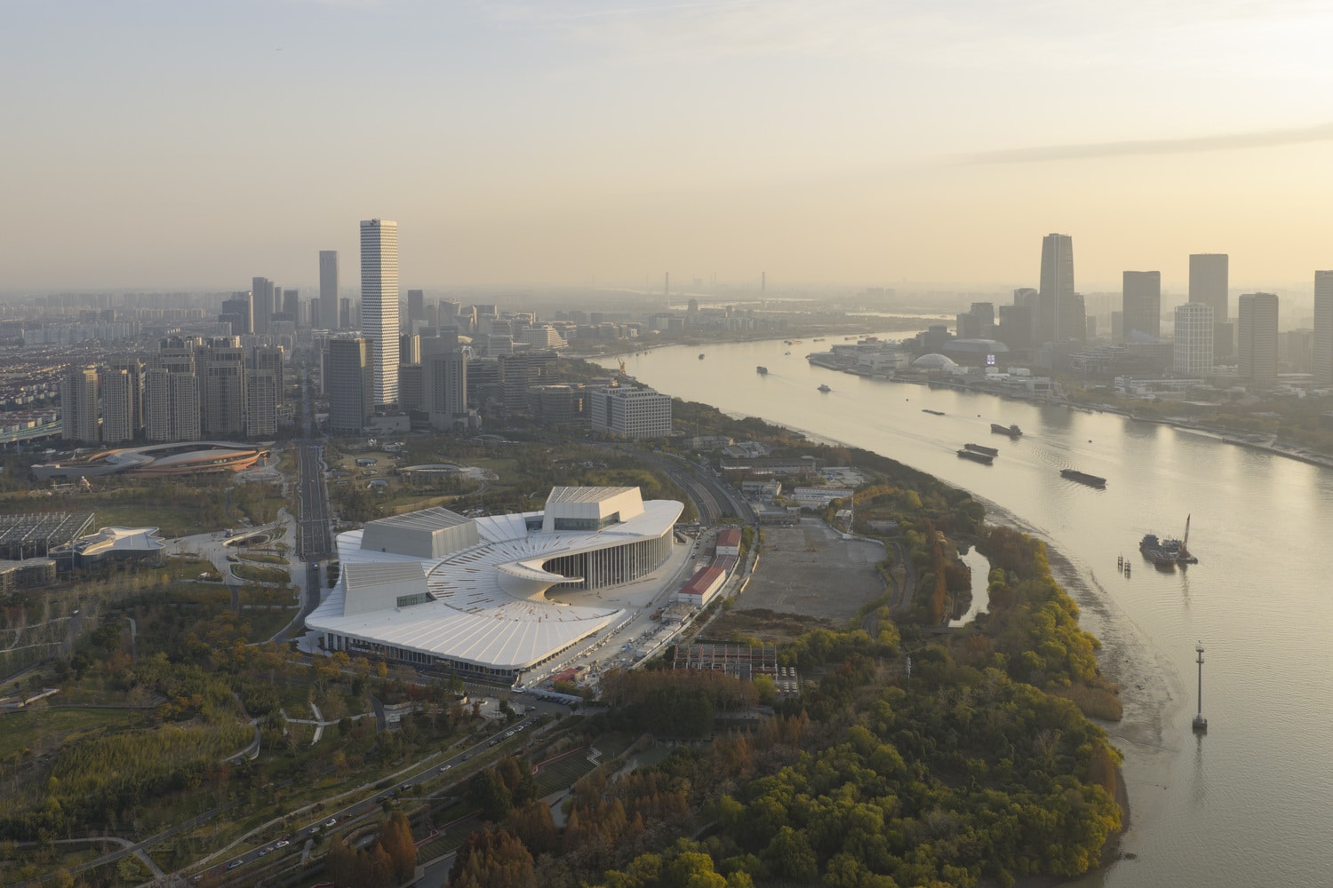 Aerial view of the Shanghai Grand Opera House featuring a white radial roof design situated along the Huangpu River at sunset.