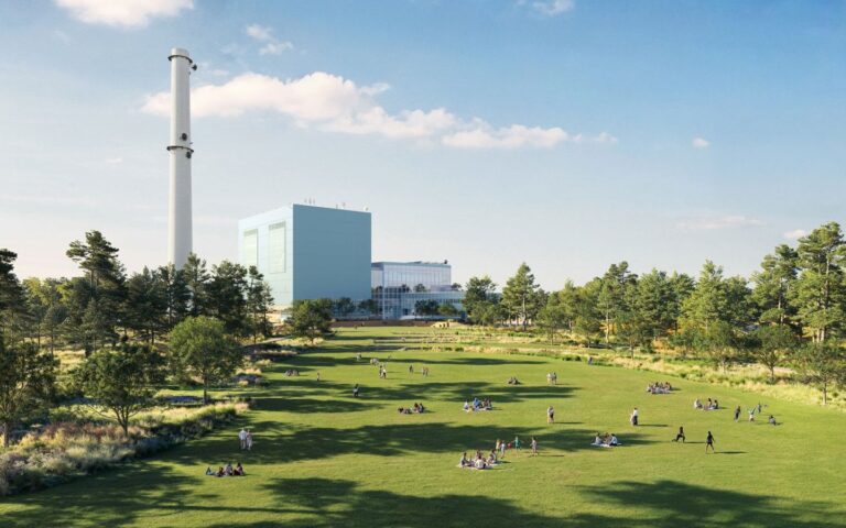 A vast green community lawn filled with people picnicking, framed by trees, with the massive blue Manresa Wilds repurposed power plant and white smokestack in the background.