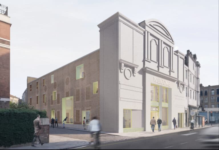 Exterior view of the Kentish Town cinema retrofit showing the preserved white Art Deco fa&ccedil;ade with a "Welcome" sign, contrasted by a new brick side elevation featuring punched windows with bright yellow frames.