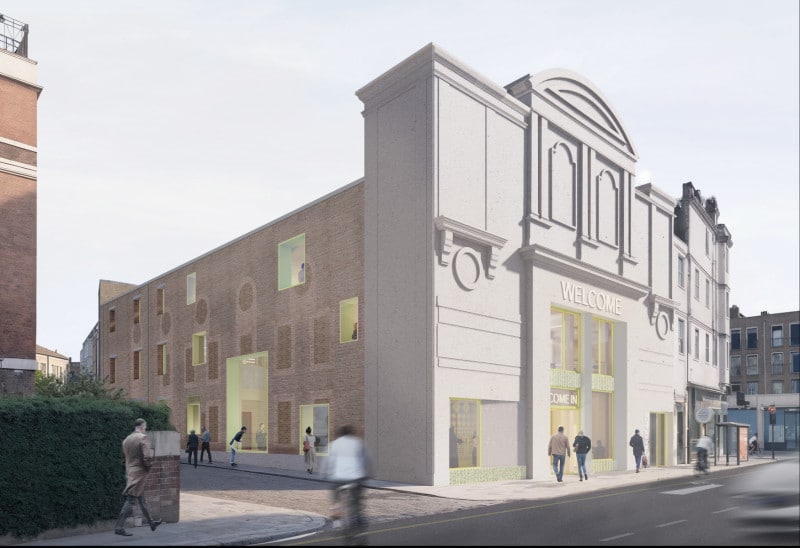 Exterior view of the Kentish Town cinema retrofit showing the preserved white Art Deco façade with a "Welcome" sign, contrasted by a new brick side elevation featuring punched windows with bright yellow frames.