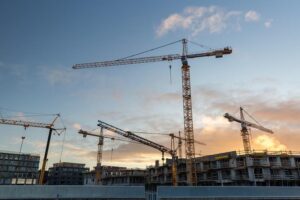 A wide shot of several tall yellow tower cranes positioned over a multi-story concrete building under construction against a blue and orange sunset sky.