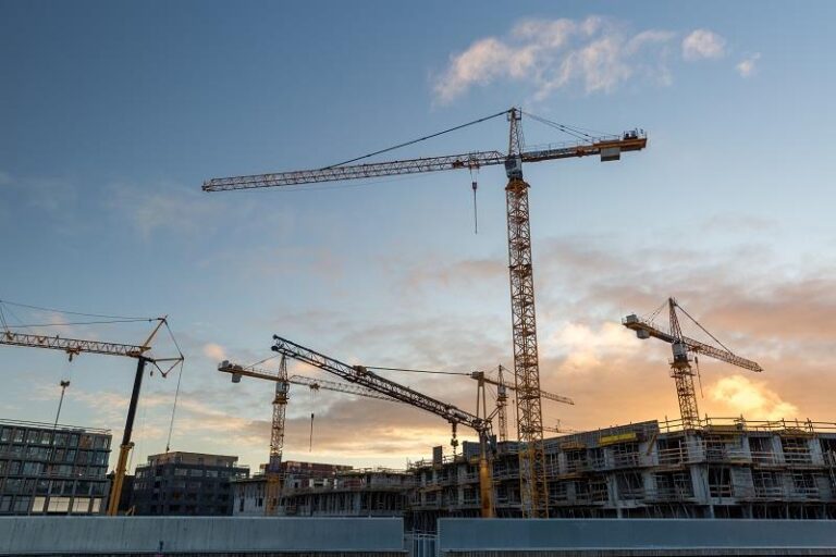 A wide shot of several tall yellow tower cranes positioned over a multi-story concrete building under construction against a blue and orange sunset sky.