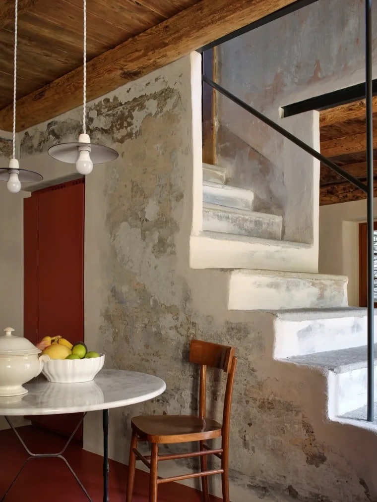 A sculptural wooden staircase framed by dark iron beams and thick stone walls, a central feature in an alpine home restoration.