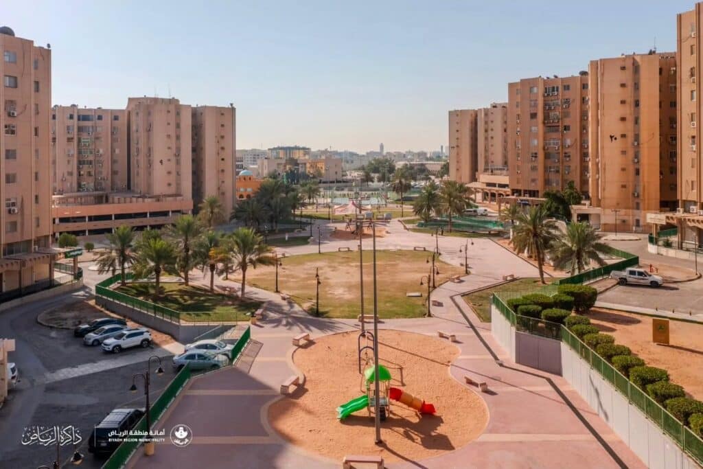 Central public park with playgrounds, palm trees, and walkways surrounded by the tall towers of the Urgent Housing complex.