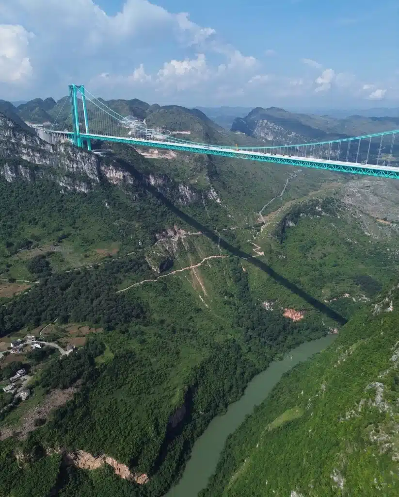 High-angle drone shot showing the Huajiang Canyon Bridge deck casting a long shadow over the rugged mountain terrain and river valley.