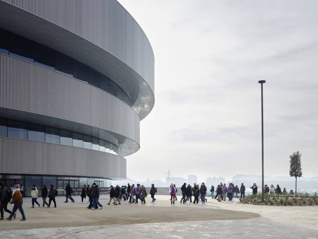 Spectators walk towards a modern, curved sports arena with a vertically slatted metallic facade on an overcast day.