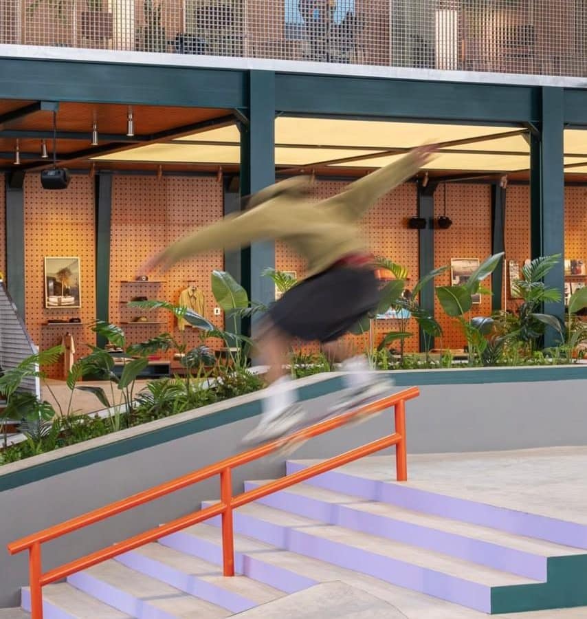 متزلج يؤدي حيلة انزلاق على سكة برتقالية فوق درج أرجواني، وتظهر في الخلفية منطقة البيع بالتجزئة بجدران خشبية ونباتات داخلية كثيفة وشرفة علوية داخل حديقة تزلج داخلية.