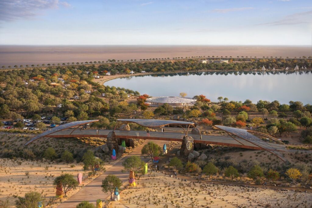A ground-level view of a paved recreational track winding through the green landscape of the Desert Oasis Project, with the calm lake and architectural pavilions in the background.