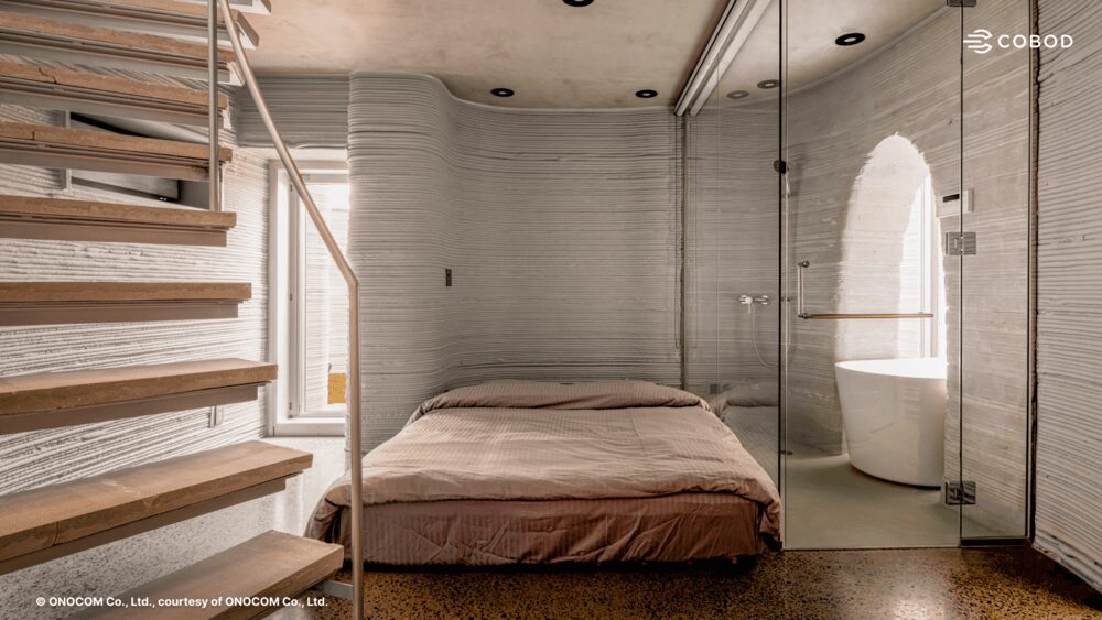 aThis image showcases the "cave-inspired" architectural volume. The horizontal striations&mdash;typical of the layer-by-layer printing process&mdash;are clearly visible on the grey concrete facade. The structure creates a striking contrast with the natural surroundings and the rigid concrete retaining wall, demonstrating the printer's ability to create free-form geometry reaching seven meters in height.aInterior bedroom space with a glass-enclosed bathroom inside a 3D-printed house, highlighting the textured layered walls and polished flooring.