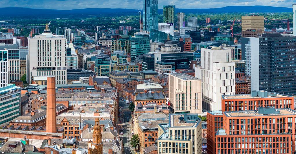 High-angle aerial view of Manchester city centre showing a dense mix of historic red-brick buildings, modern glass skyscrapers, and industrial chimneys under a bright, partly cloudy sky.