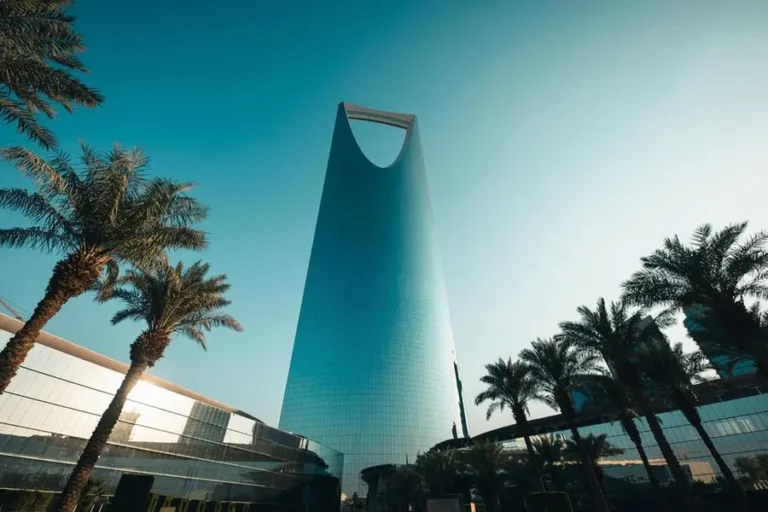 Low-angle shot of the glass-clad Kingdom Centre skyscraper in Riyadh, Saudi Arabia, framed by palm trees against a bright, clear teal sky.