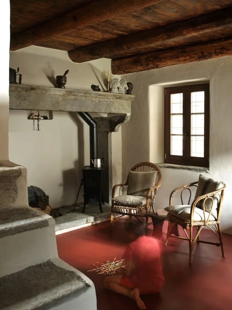 A cozy living area in a renovated alpine home featuring a large stone fireplace, a modern wood stove, and bold red flooring.