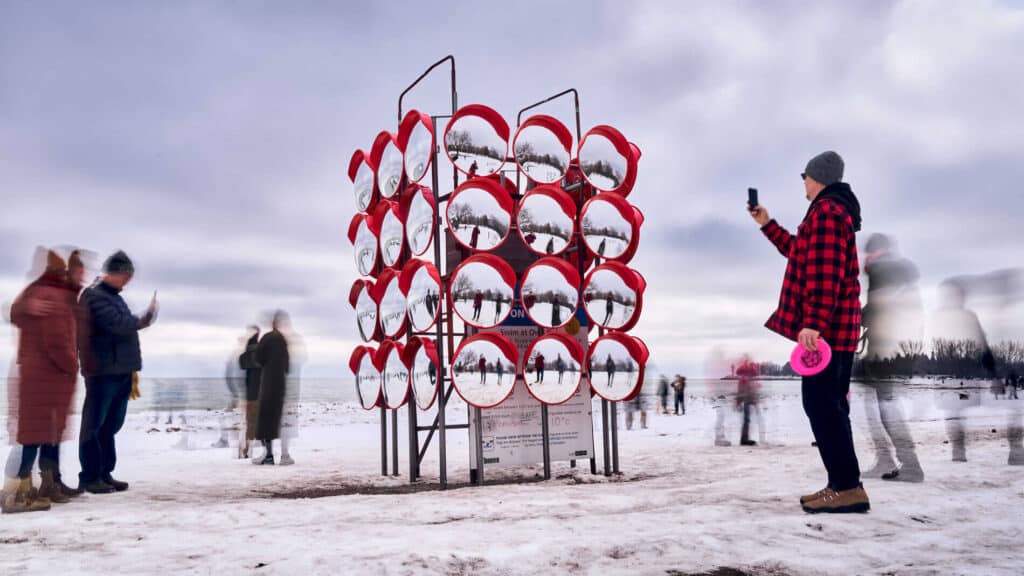 Steel tower structure holding a grid of red-backed convex mirrors reflecting a snowy park scene.