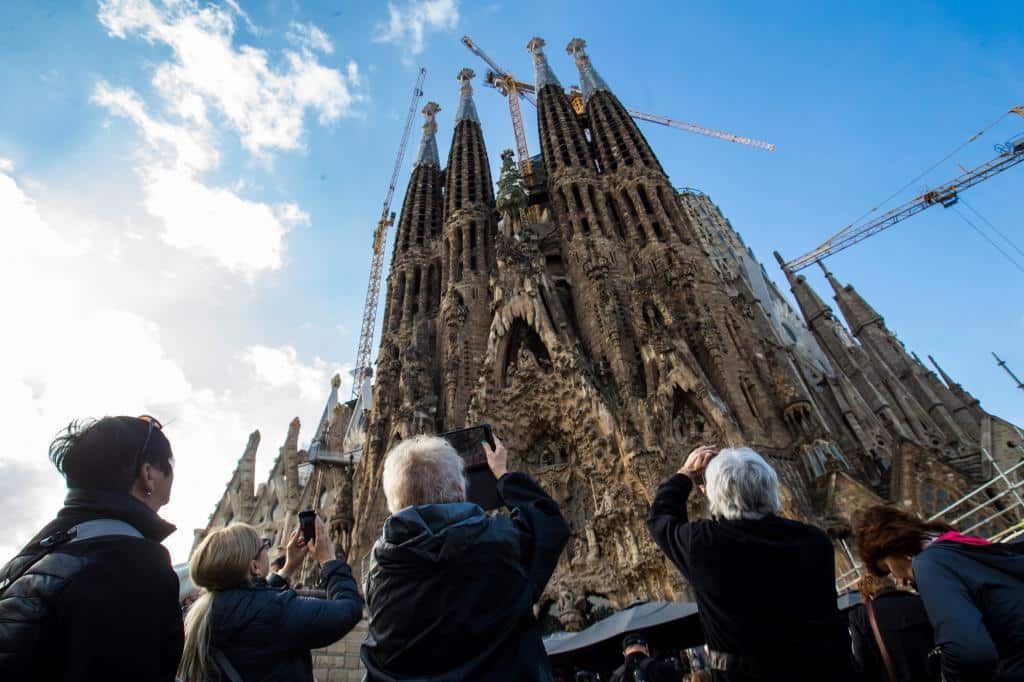 Tourists gathered at street level photographing the massive intricate facades and construction cranes of the basilica against a cloudy sky.