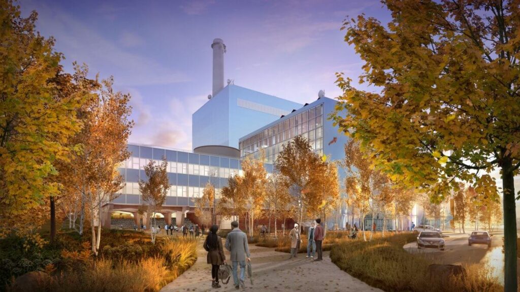 Pedestrians walking on a nature trail lined with autumn trees near the renovated glass-facade administration building and the towering smokestack.