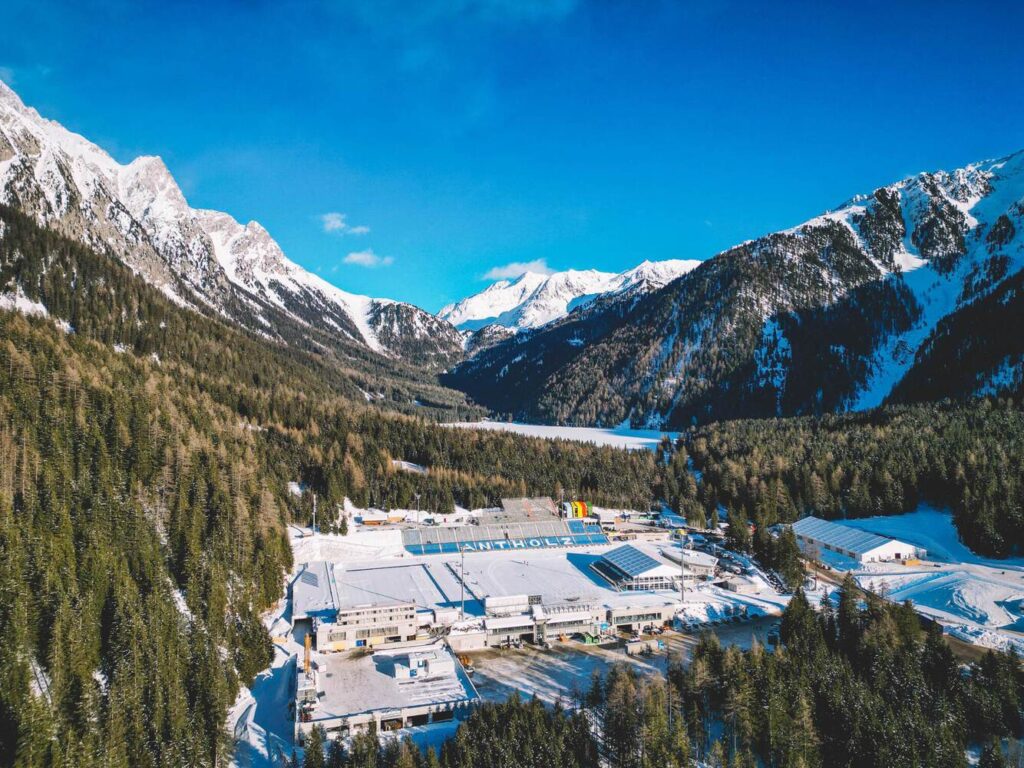 The Antholz Biathlon Stadium, a key venue for the Milano Cortina 2026 Games, is seen from above, nestled in a snowy Alpine valley surrounded by pine forests and mountains.