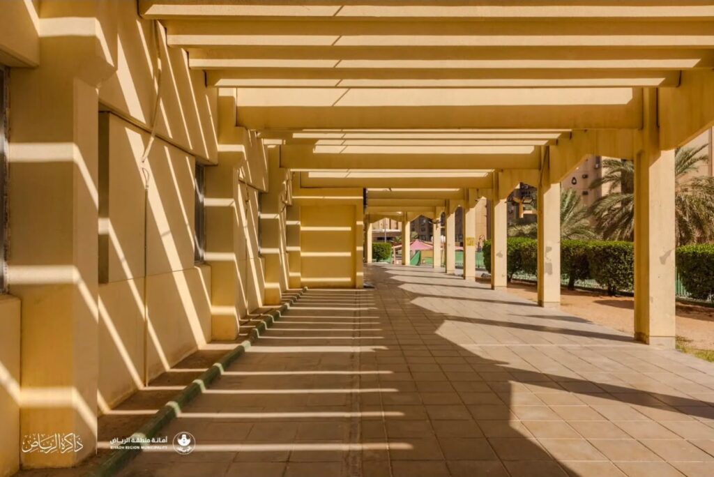 Long shaded concrete walkway with rhythmic overhead beams casting geometric shadows on the beige walls and tiled floor