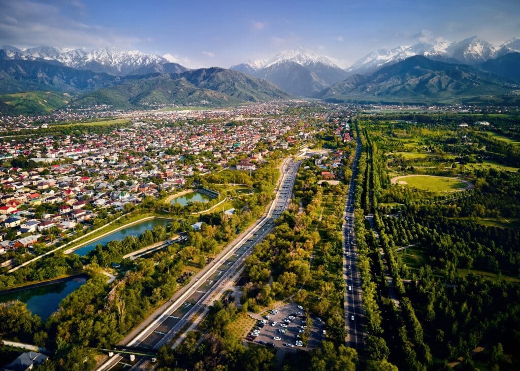 Drone shot of a straight water channel dividing residential zones and green spaces, leading the eye toward snow-capped mountains and Almaty architecture.