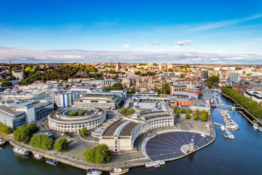 An aerial drone shot of the Bristol Harbourside Urban Regeneration, showcasing the curved Millennium Square, We The Curious science center, the waterfront amphitheater, and the city skyline.