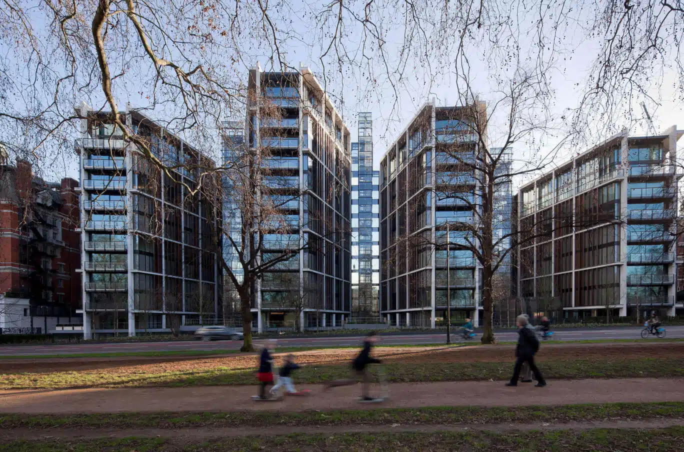A wide shot of the One Hyde Park residential complex in London, featuring modern glass and steel towers seen through bare winter trees, with people and children blurred in motion on a park path in the foreground.