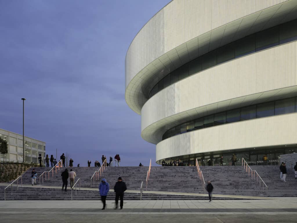 At dusk, crowds gather on the grand illuminated stairs of a new, curvilinear arena built for the Milano Cortina 2026 games.