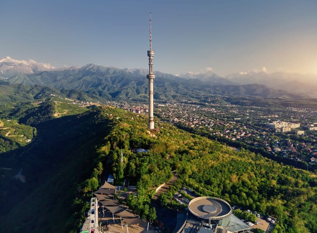 The Almaty Television Tower rising from the lush greenery of Kok Tobe hill, illuminated by golden hour sunlight.