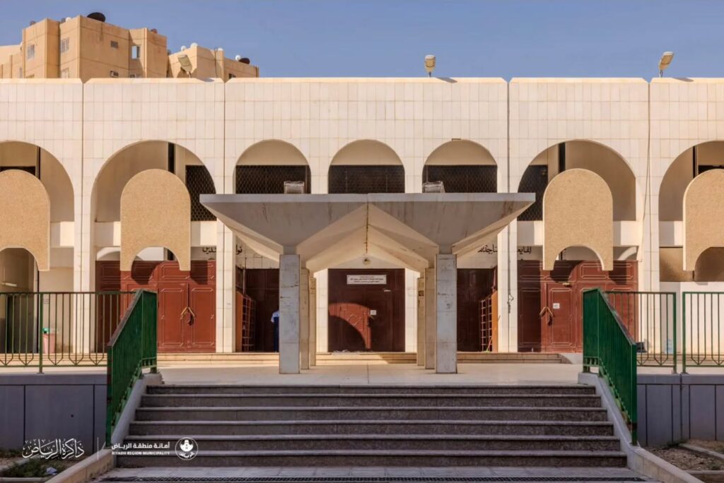 واجهة أمامية لمبنى مجتمعي تضم سلسلة من الأقواس ومظلة خرسانية مركزية عند المدخل وسلالم ذات درابزين أخضر.