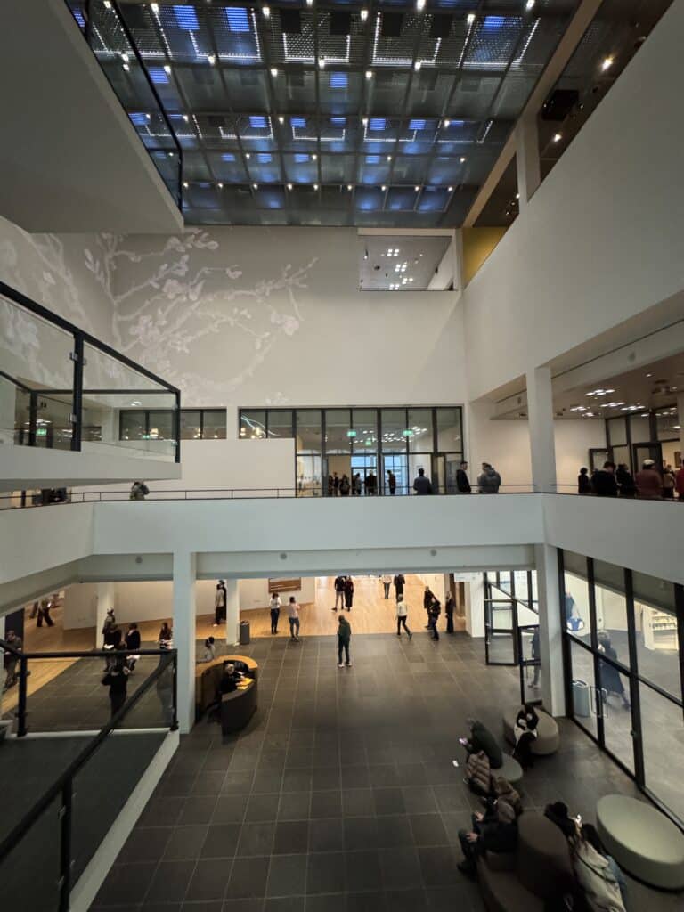 A spacious, multi-level modern museum atrium featuring a translucent glass ceiling, minimalist white walls, and visitors exploring the interior spaces.