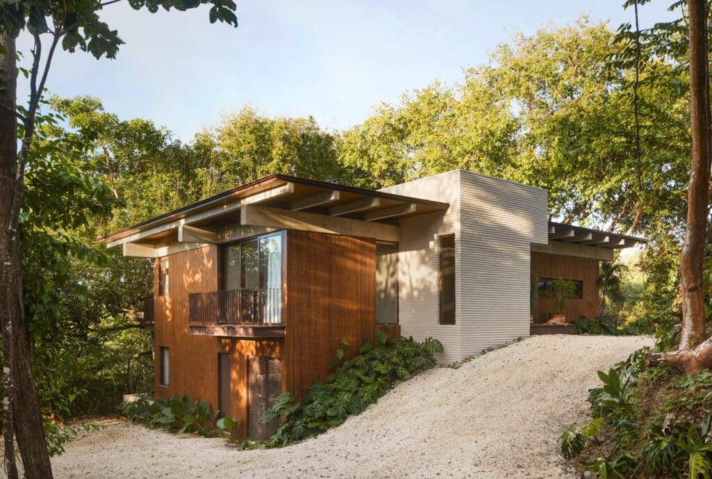 A two-story modern house with warm wood paneling and a textured light-grey concrete core, nestled into a lush green forest hillside with a gravel driveway.