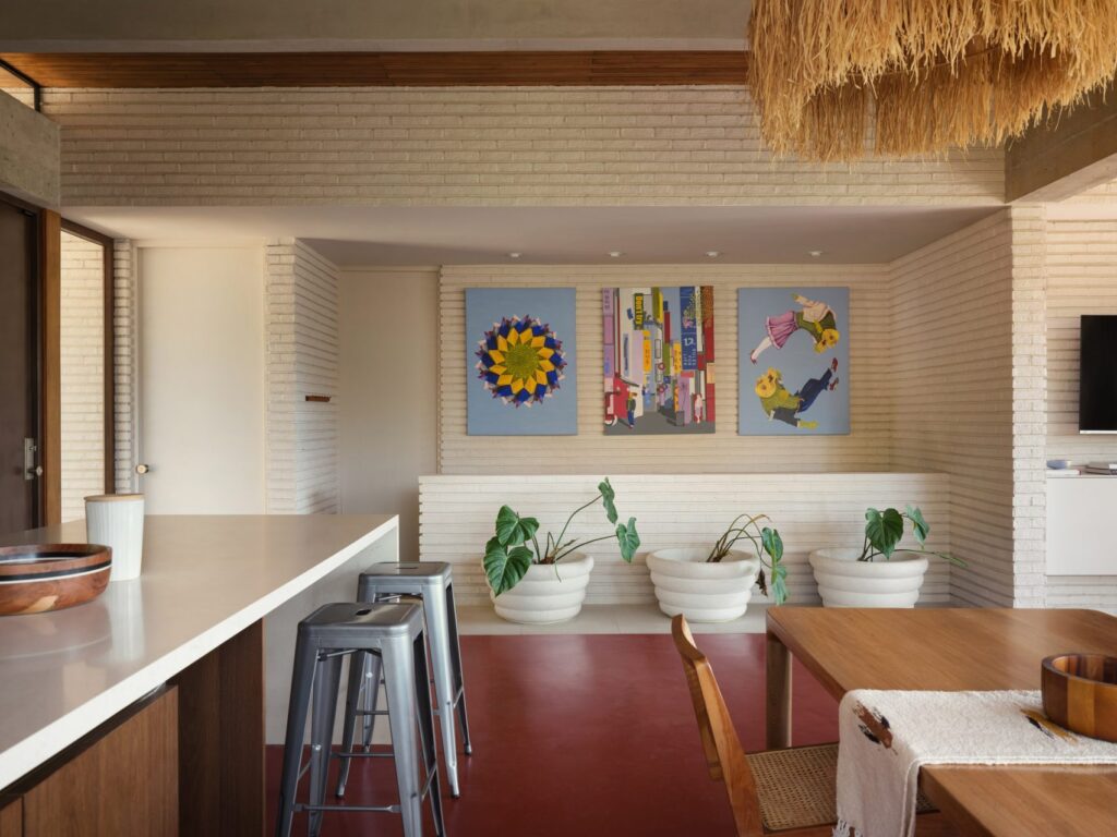 A modern open-plan dining and kitchen area featuring white brick walls, a deep red floor, and three colorful contemporary paintings.