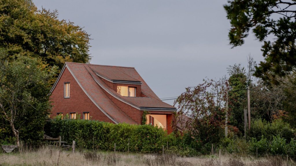 Modern house with a sweeping red clay tile roof and walls, partially hidden by green hedges under a twilight sky.