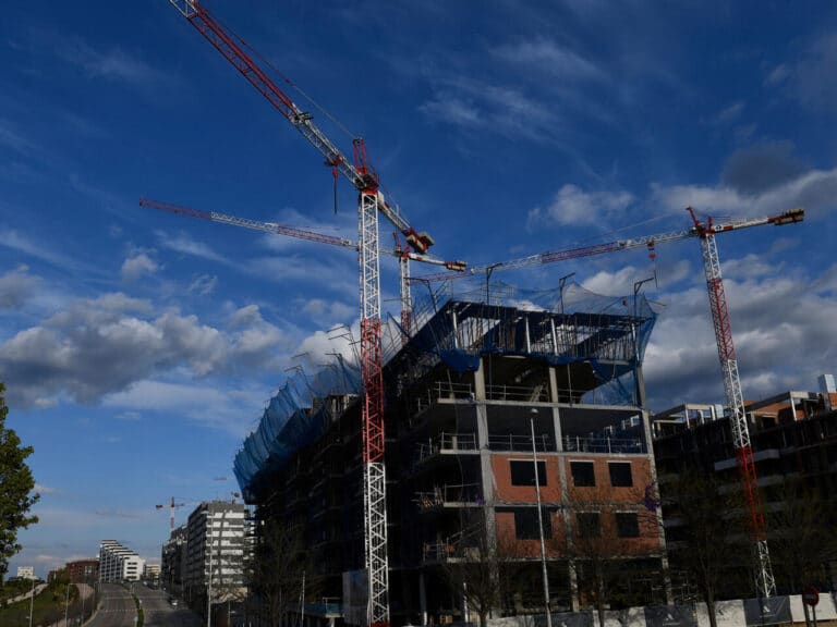 A high-angle shot of a multi-story building under construction, surrounded by three large red and white tower cranes against a blue sky with scattered clouds.